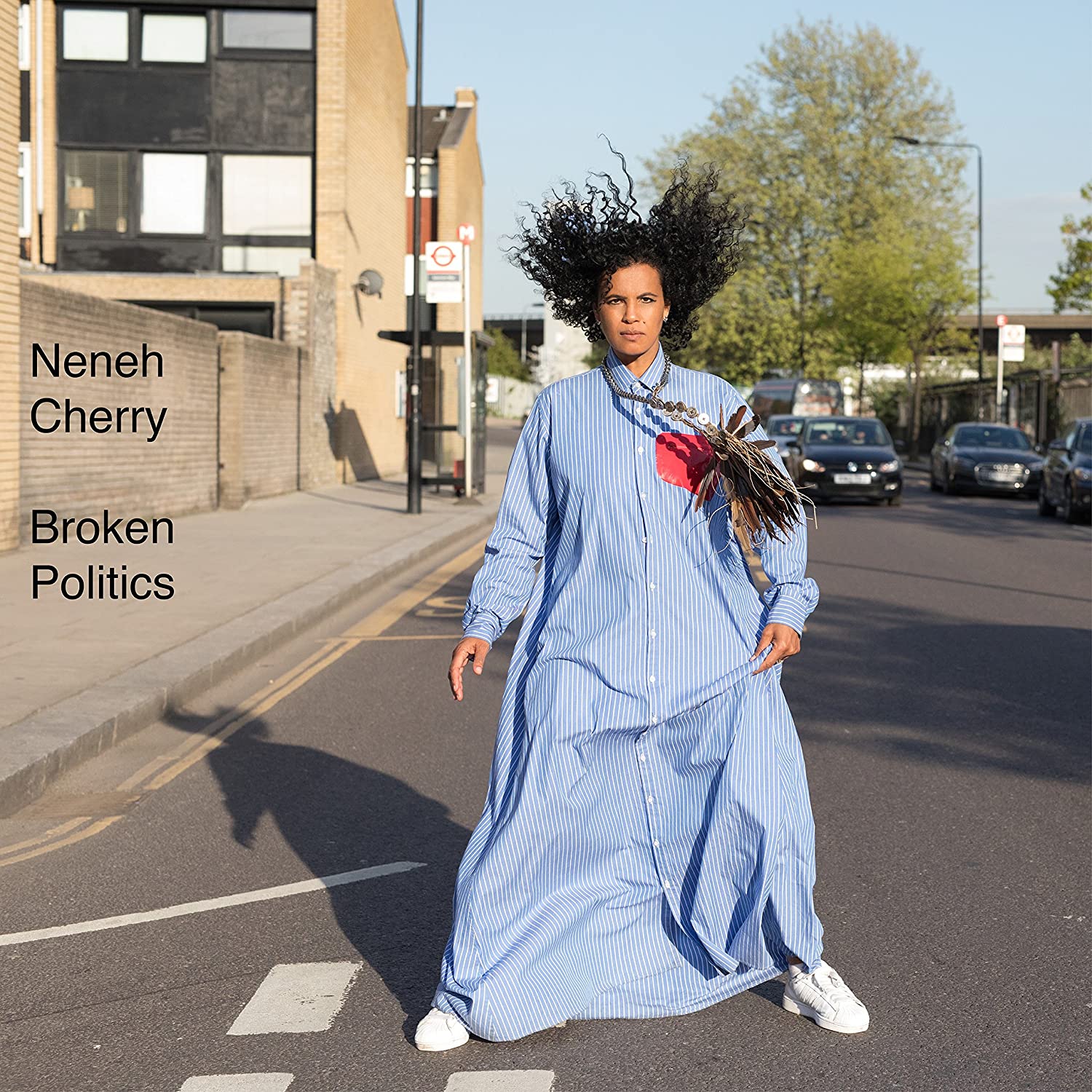 the album cover for broken politics. pictured, cherry is in a windswept floor length pinstred blue gown and white sneakers. their hair floats straight into the air. behind them, a  residential building. trees and cars litter the street