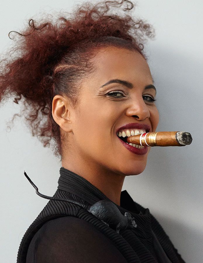 Cherry is pictured smoking a cigar with a confident smile and looking toward the viewer. On their shoulder is a black toy rat.